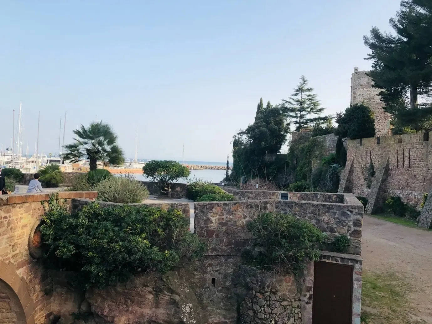Vue d'une forteresse en pierre avec des murs anciens, des plantes et des arbres, donnant sur un port avec des bateaux