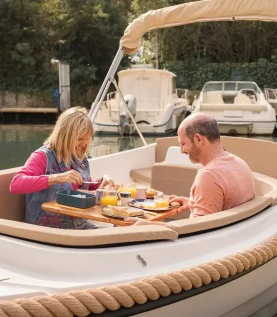 Un couple prend un repas sur un bateau amarré dans un canal, avec des plats et des boissons disposés sur une table entre eux