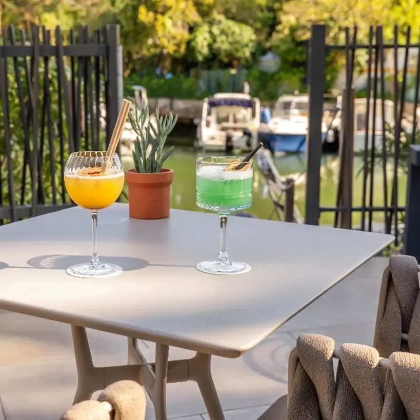 Deux verres de cocktails colorés sur une table en terrasse près d'un canal avec des bateaux, entourés de plantes et de chaises