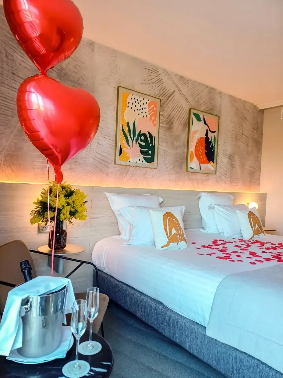 Chambre avec un lit recouvert de pétales rouges, deux ballons en forme de cœur, un vase avec des fleurs, une bouteille de champagne dans un seau à glace et deux flûtes à champagne sur une table