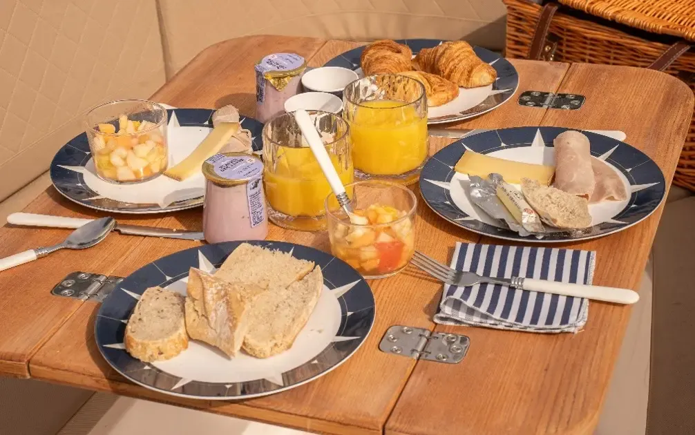 Table de petit-déjeuner avec des plats contenant des croissants, des fruits, du fromage, des charcuteries, des tartines, de la confiture, du jus d'orange et des couverts