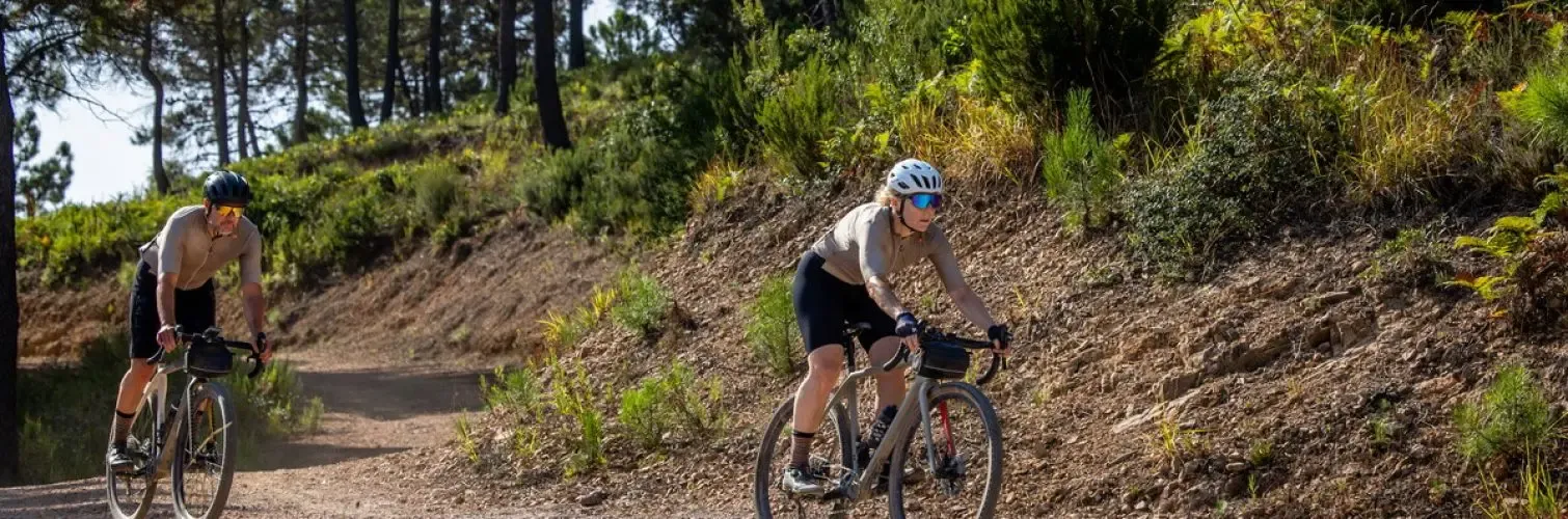 Deux cyclistes portent des casques et des lunettes de soleil, ils roulent sur un chemin de terre en forêt