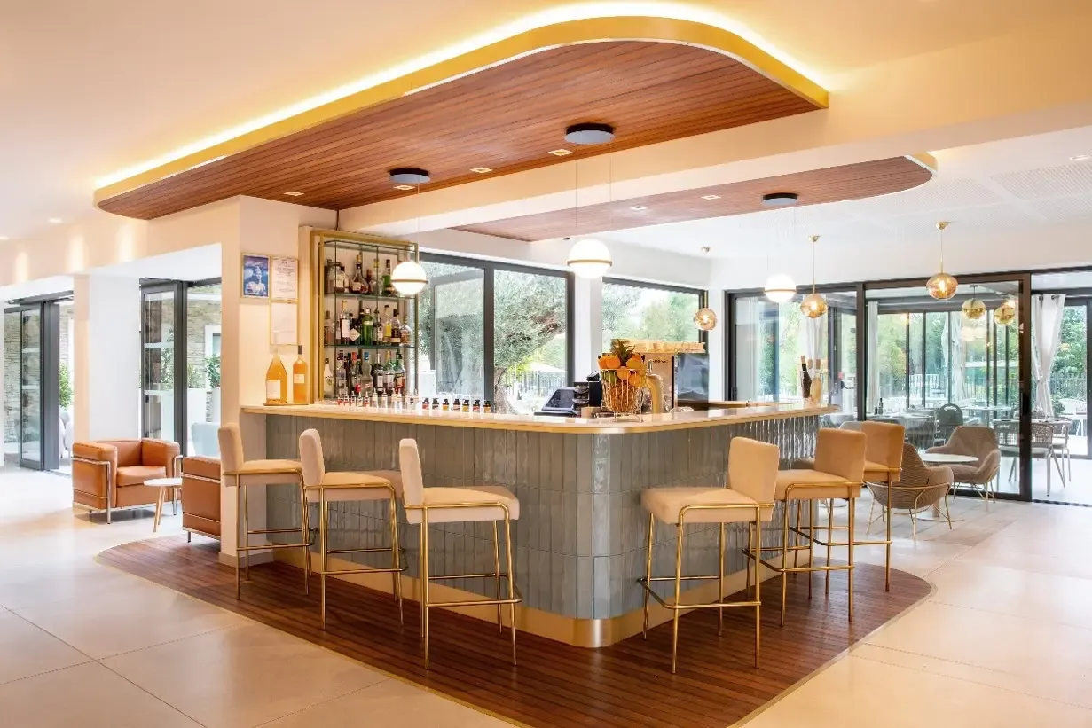 Bar moderne avec comptoir en marbre gris et tabourets en cuir beige, éclairé par des suspensions dorées, entouré de grandes baies vitrées