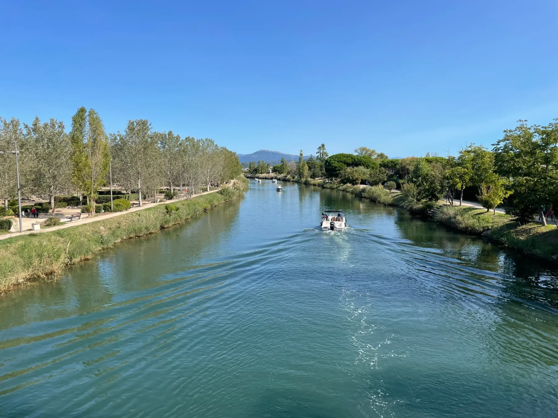 Un bateau navigue sur une rivière bordée d'arbres et de sentiers de promenade sous un ciel bleu