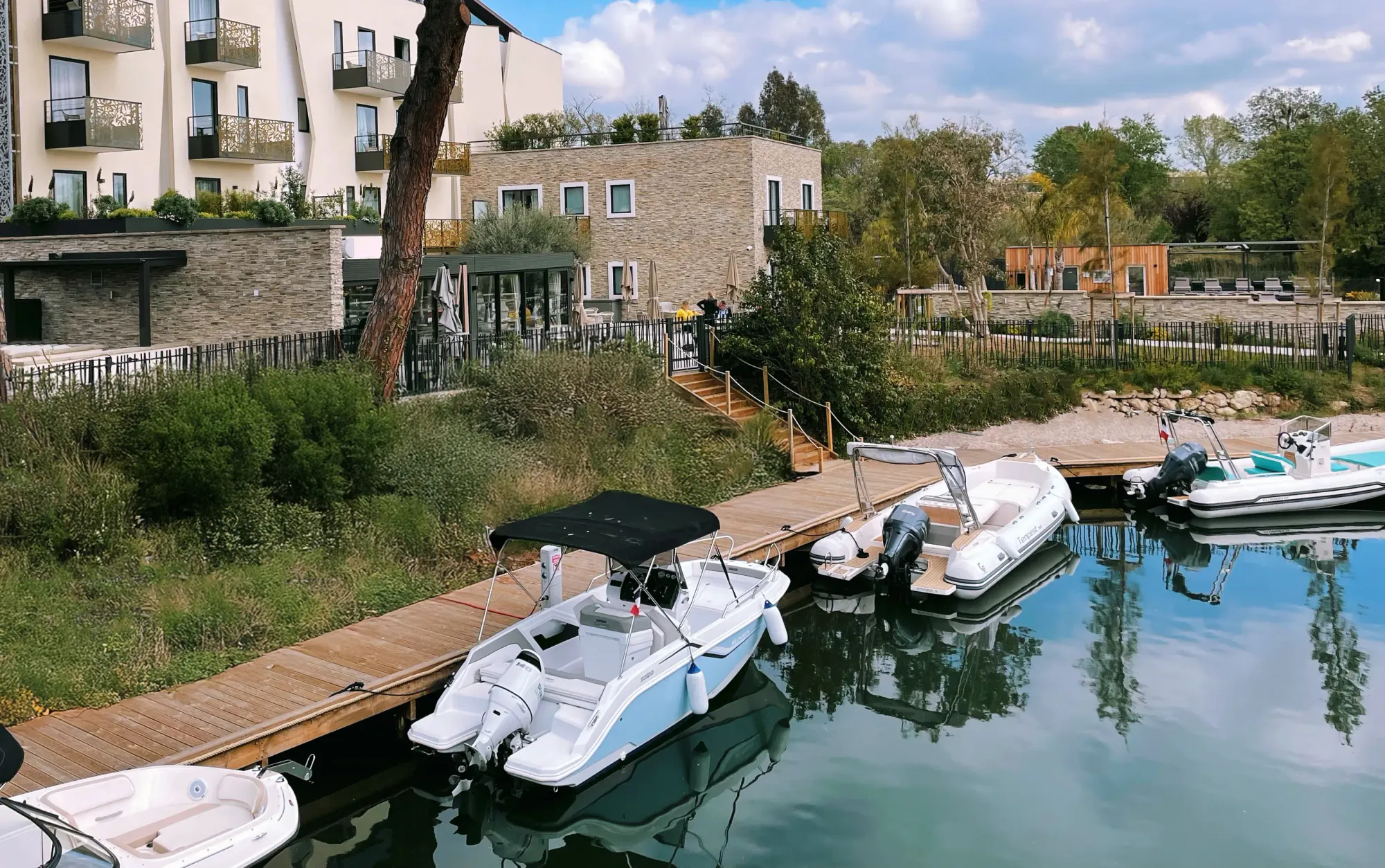 Quais en bois avec plusieurs petits bateaux à moteur amarrés devant des bâtiments résidentiels modernes entourés de végétation