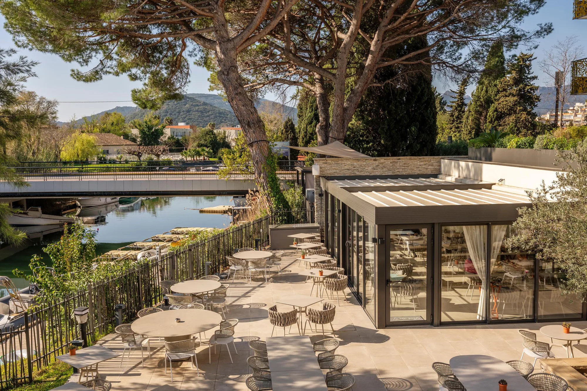Terrasse de café avec tables et chaises en plein air, bordée d'arbres et d'un plan d'eau, située près d'un bâtiment moderne aux grandes baies vitrées, avec des montagnes en arrière-plan