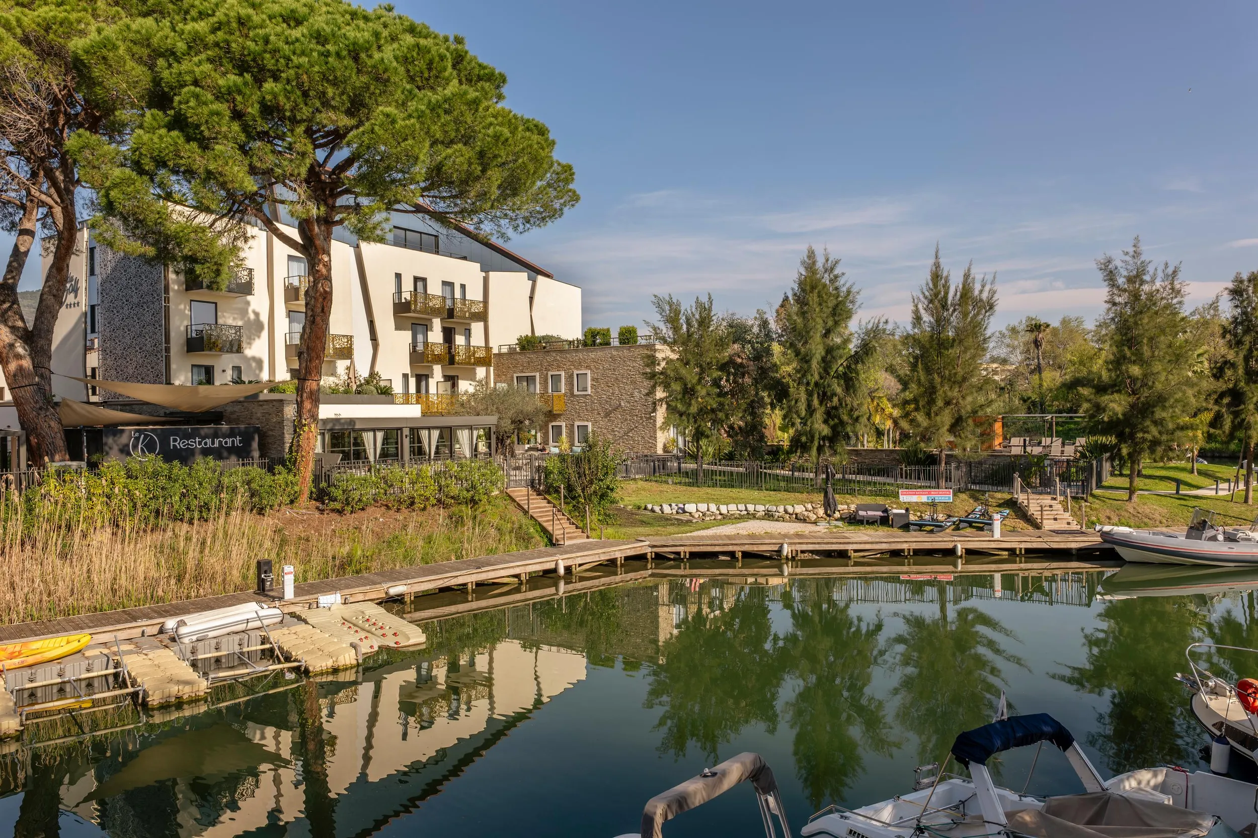 Un bâtiment avec un restaurant et des balcons surplombe un canal bordé d'arbres et de bateaux amarrés