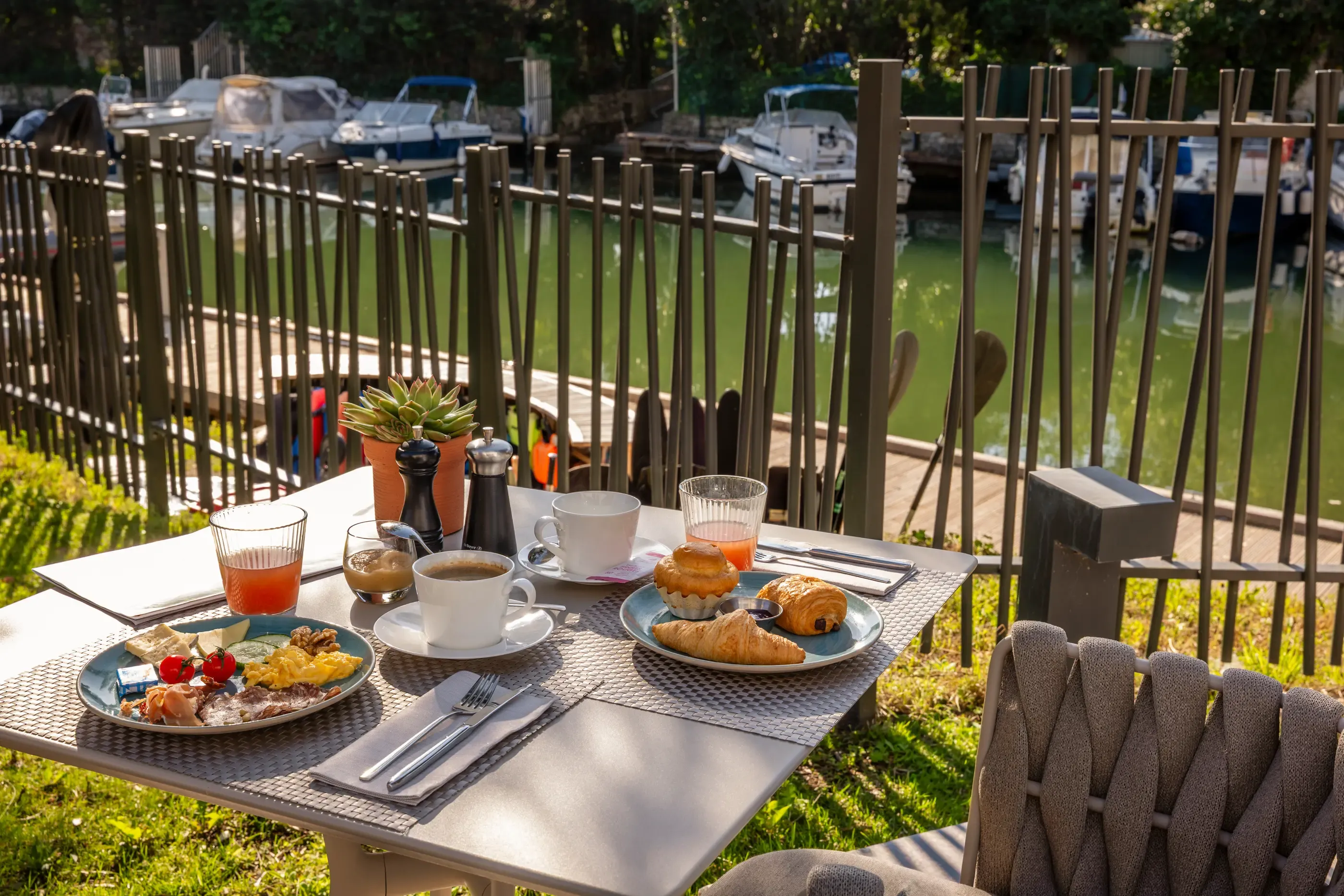 Table en extérieur avec petit-déjeuner composé de croissants, pain, confiture, fruits et boissons, vue sur un canal avec des bateaux