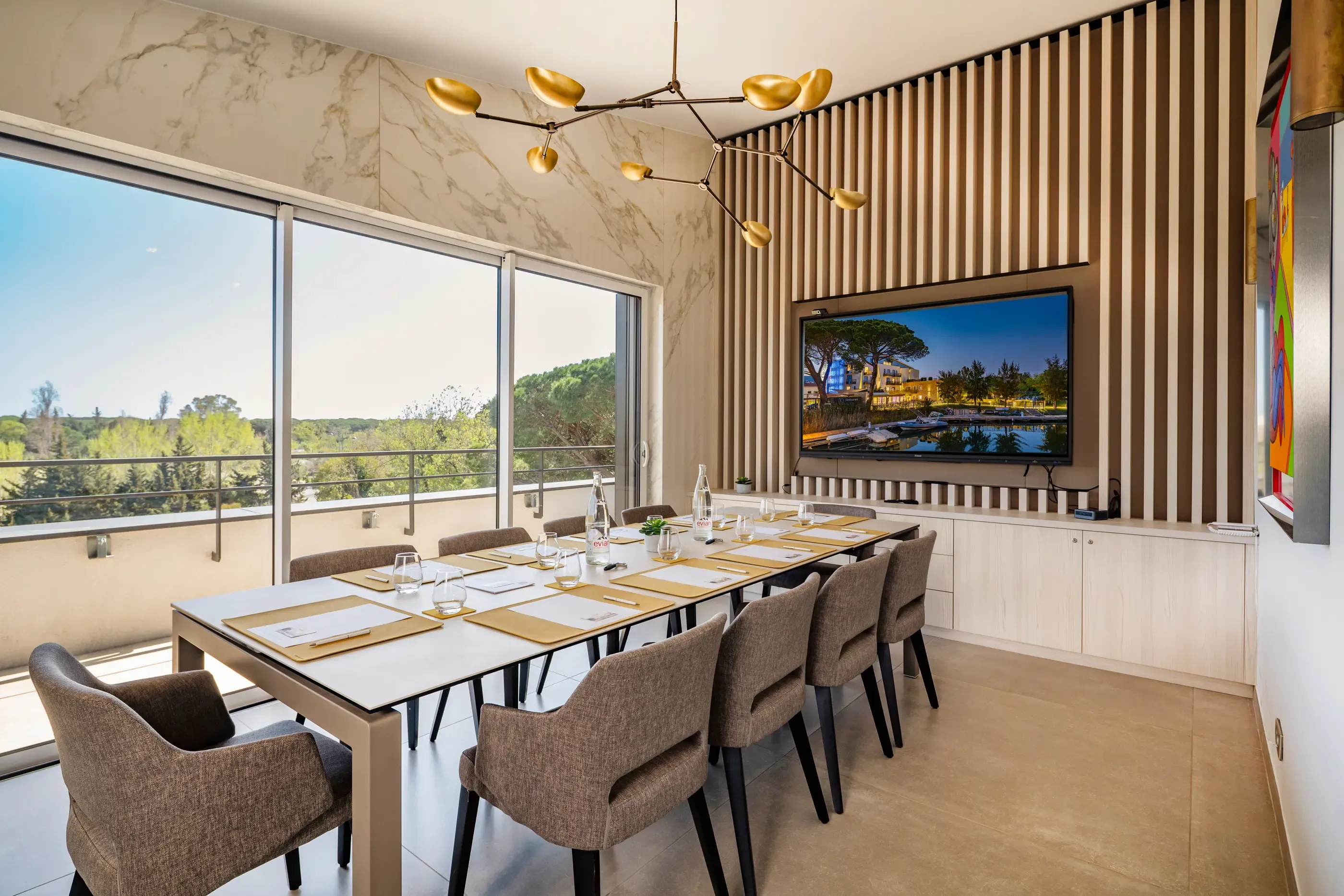 Salle de réunion avec une table, des chaises, une télévision affichant un paysage et de grandes fenêtres offrant une vue sur l'extérieur
