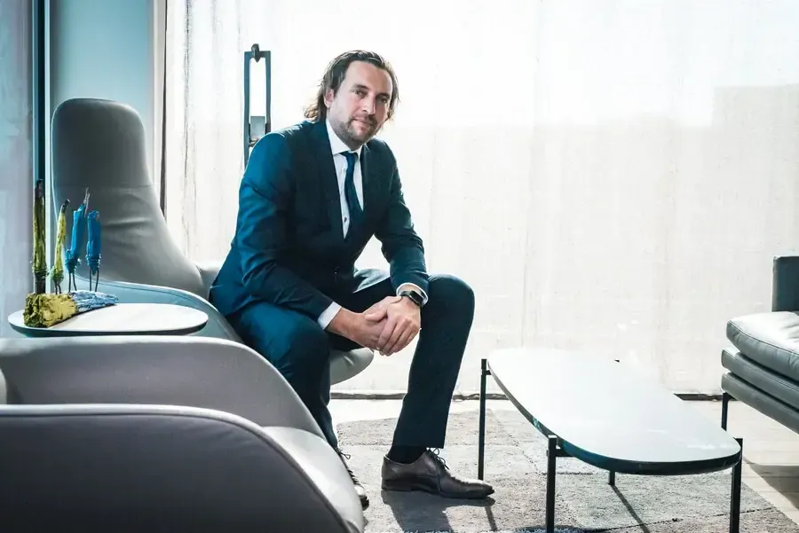Un homme en costume bleu est assis sur un canapé gris dans un espace moderne avec une table basse et des plantes décoratives