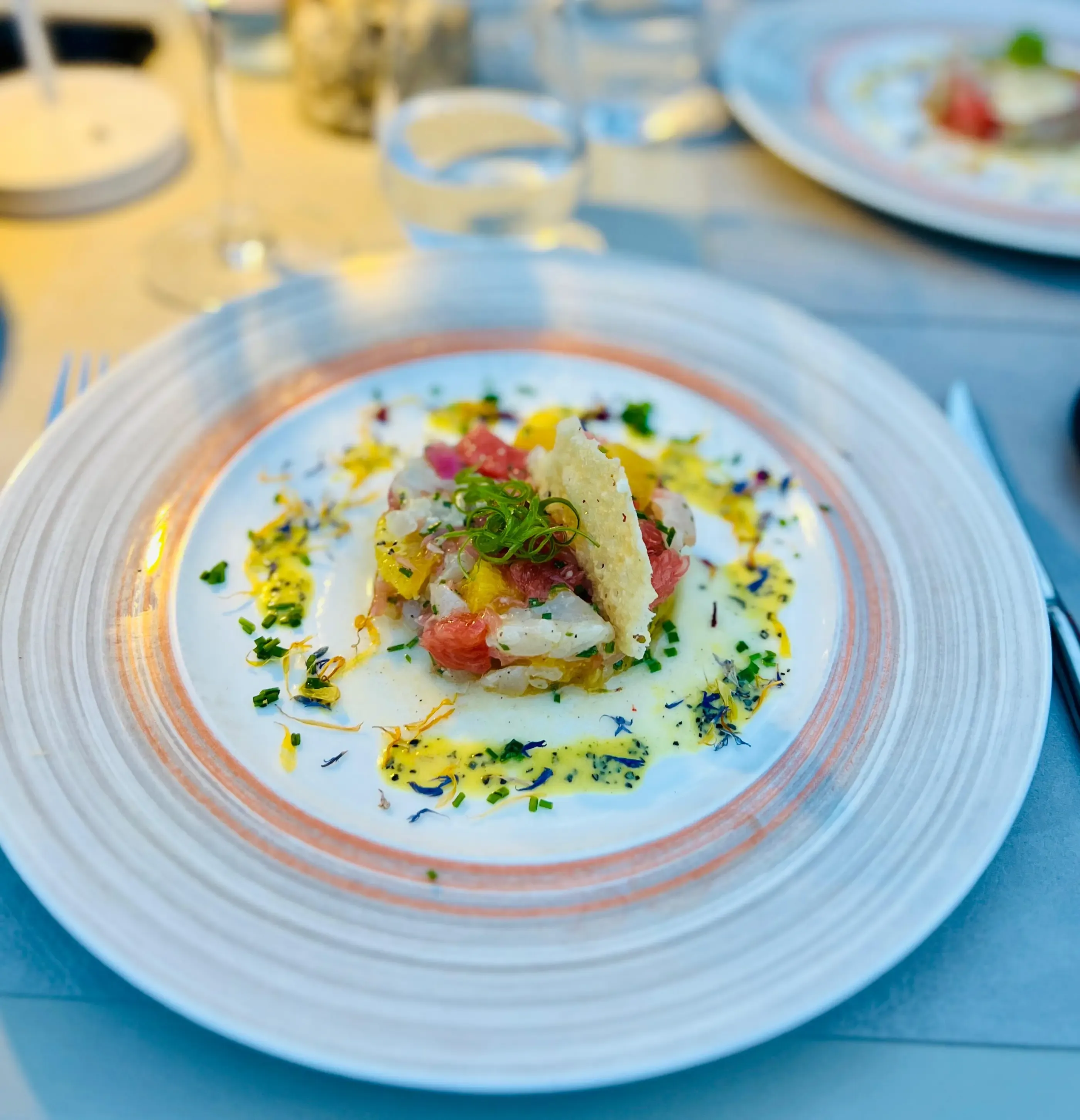 Assiette avec une préparation culinaire composée de tarte fine, de poisson cru et de garnitures variées, posée sur une table bleue