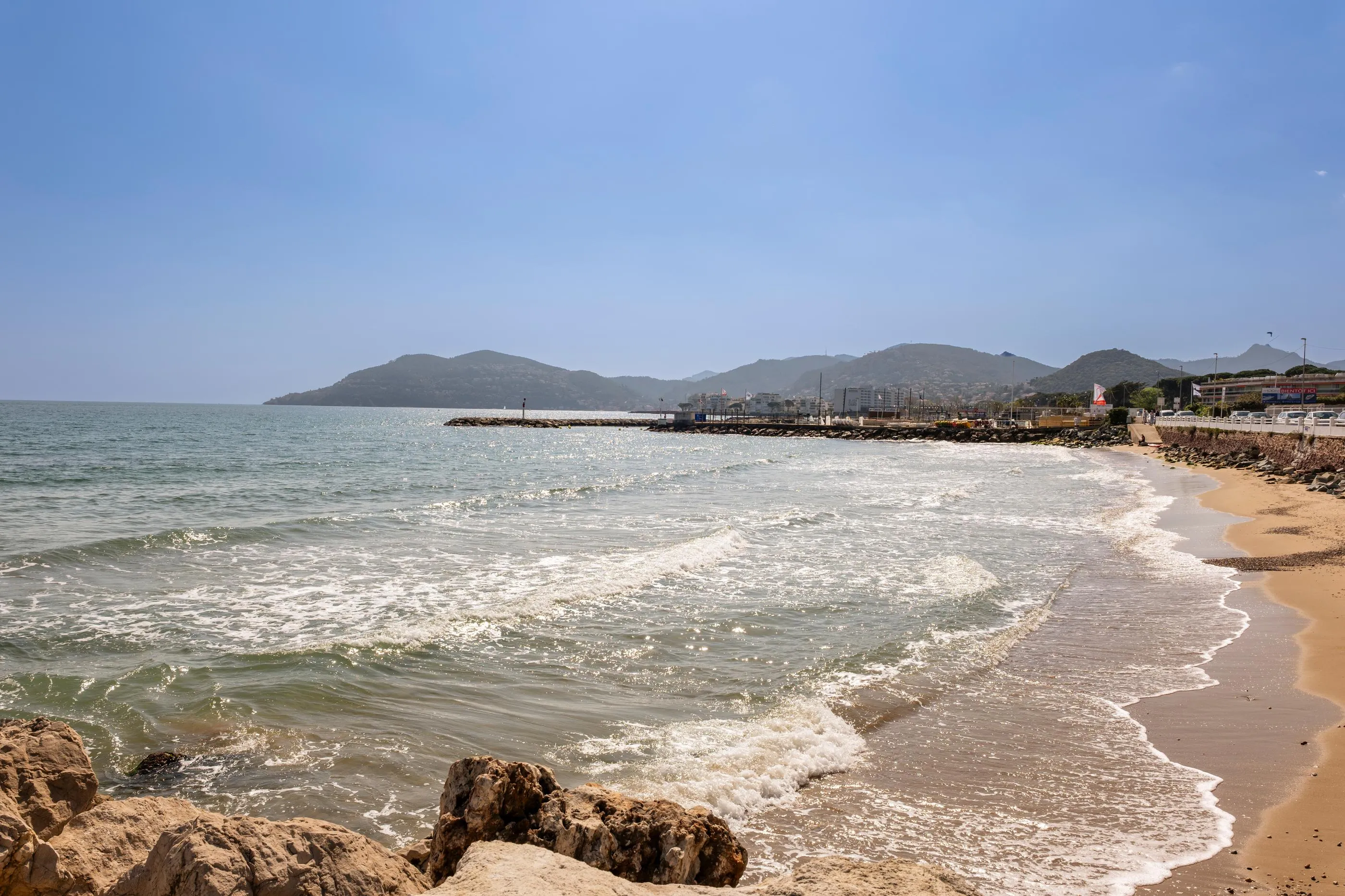 Vaguelette sur une plage de sable avec des rochers, bordée par une mer calme et des collines en arrière-plan