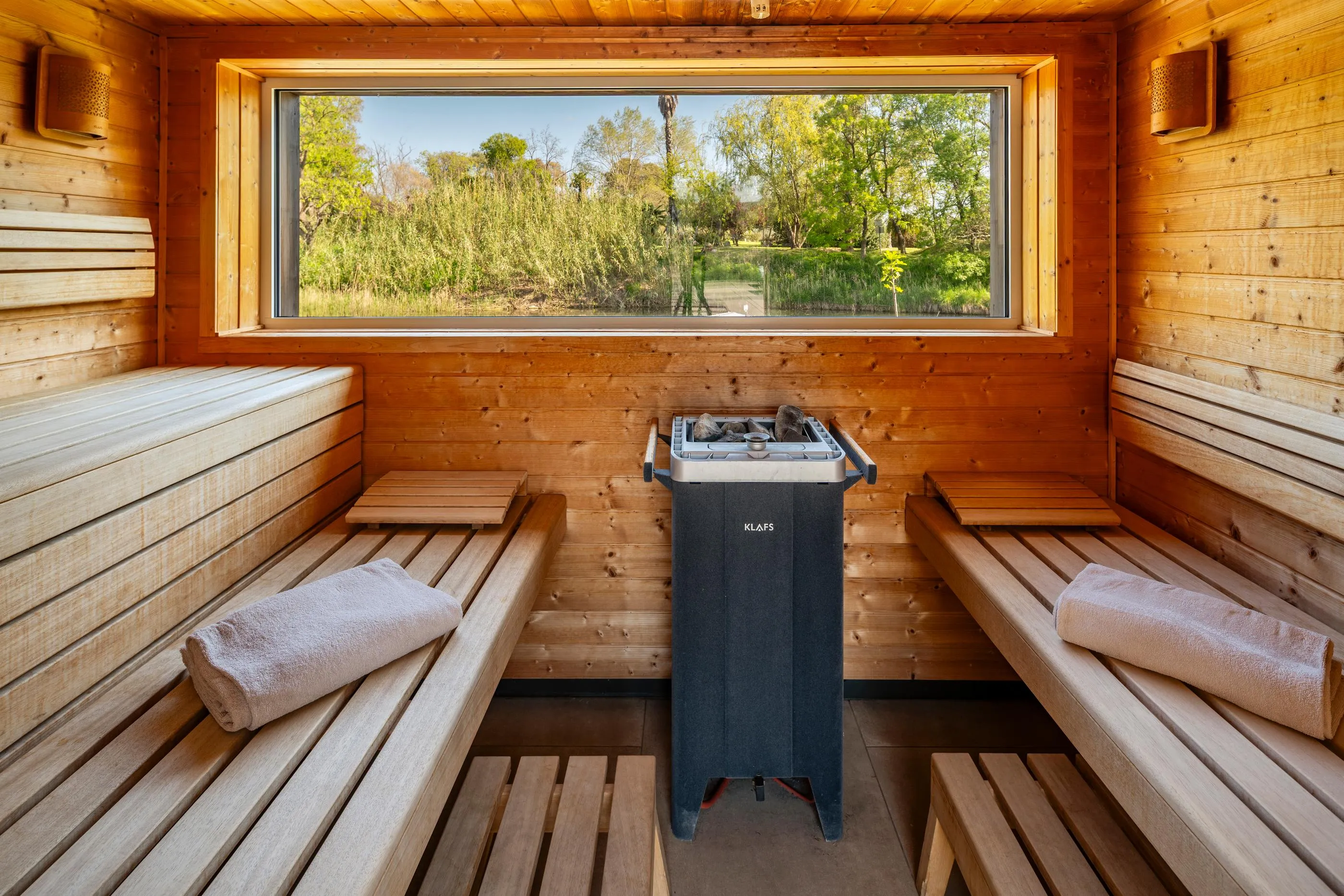 Sauna en bois avec bancs, serviettes et un poêle chauffant, vue sur un paysage extérieur à travers une fenêtre