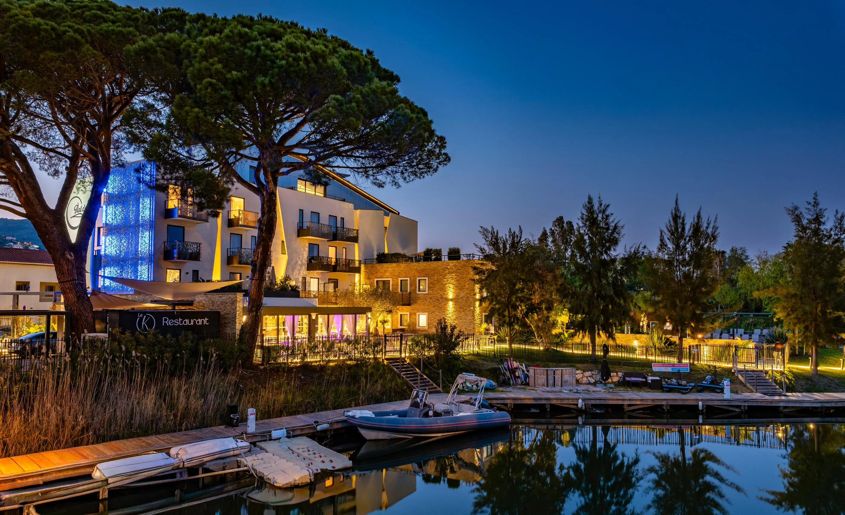 Un bâtiment moderne éclairé avec un restaurant au rez-de-chaussée, entouré d'arbres et situé près d'un plan d'eau avec des bateaux amarrés