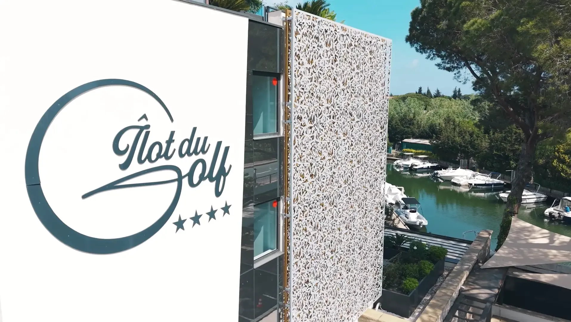 Panneau avec le logo et le nom "Îlot du Golf" à quatre étoiles près d'un plan d'eau avec des bateaux
