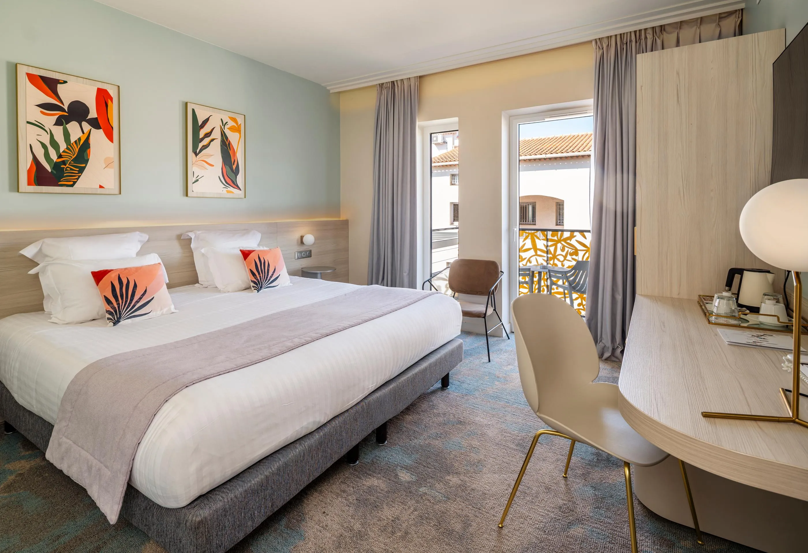 Chambre d'hôtel avec un lit double, une table de chevet, une chaise et une porte-fenêtre donnant sur un balcon avec une vue sur des toits