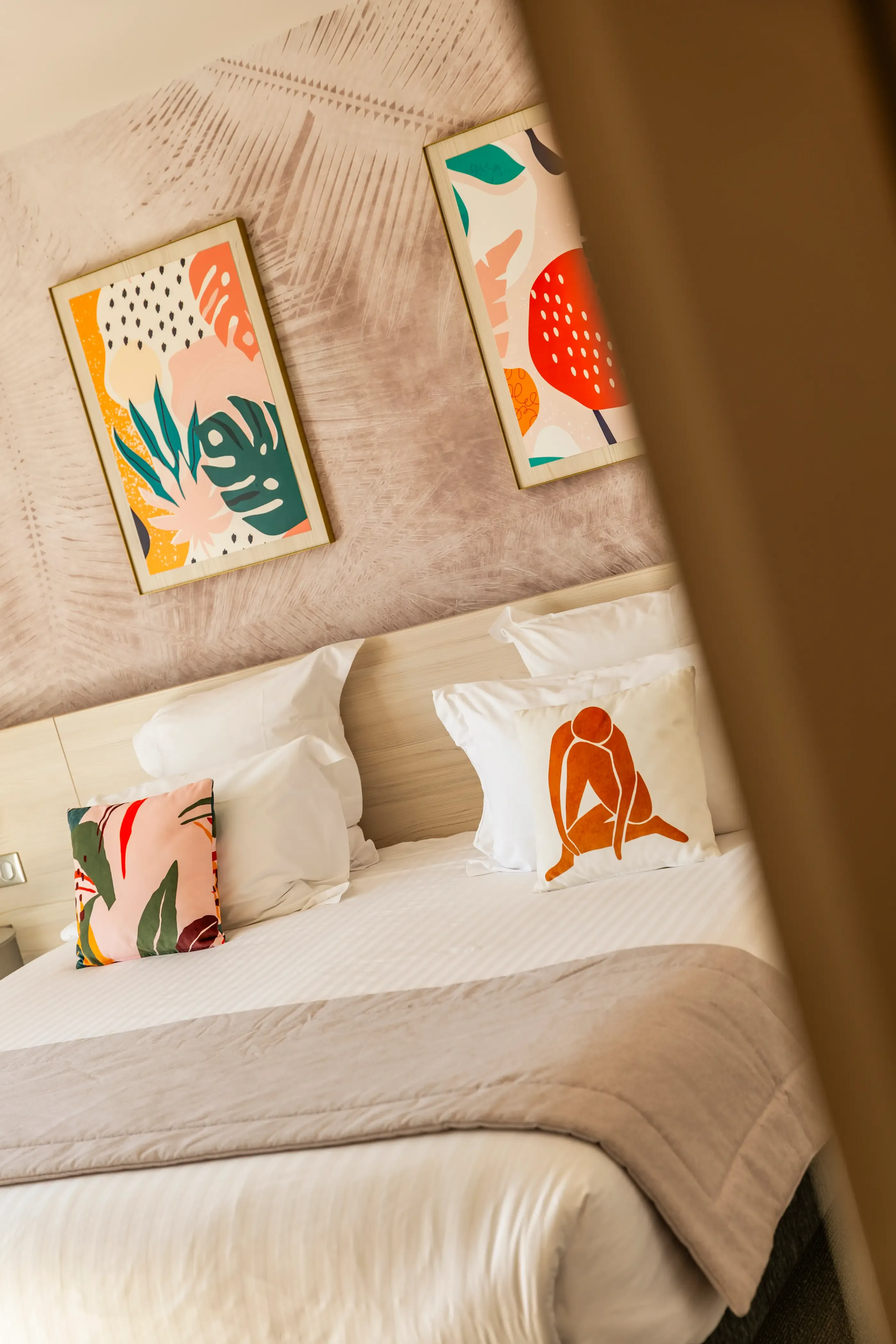 Un lit avec une tête de lit en bois clair, des coussins colorés et deux tableaux abstraits accrochés au mur