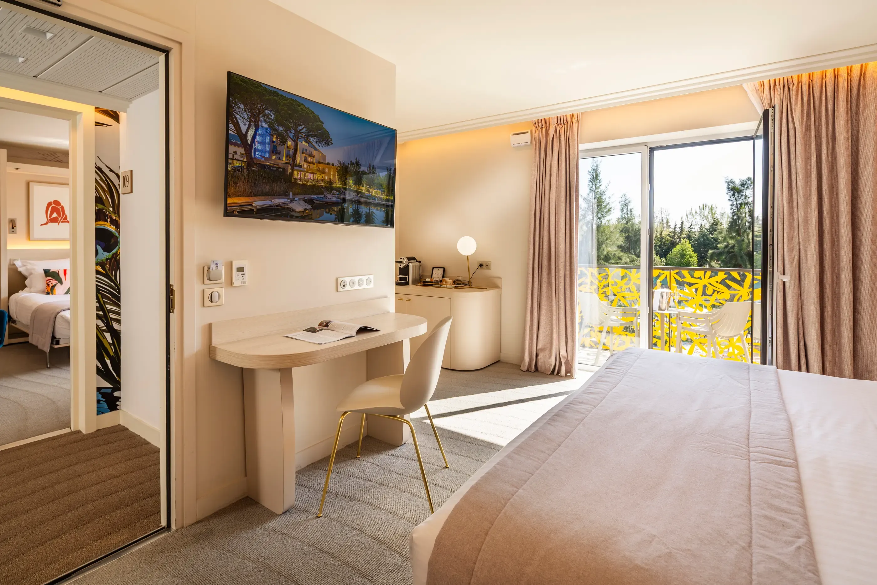 Chambre d'hôtel avec un lit, un bureau, une télévision et une porte-fenêtre donnant sur un balcon avec vue sur des arbres