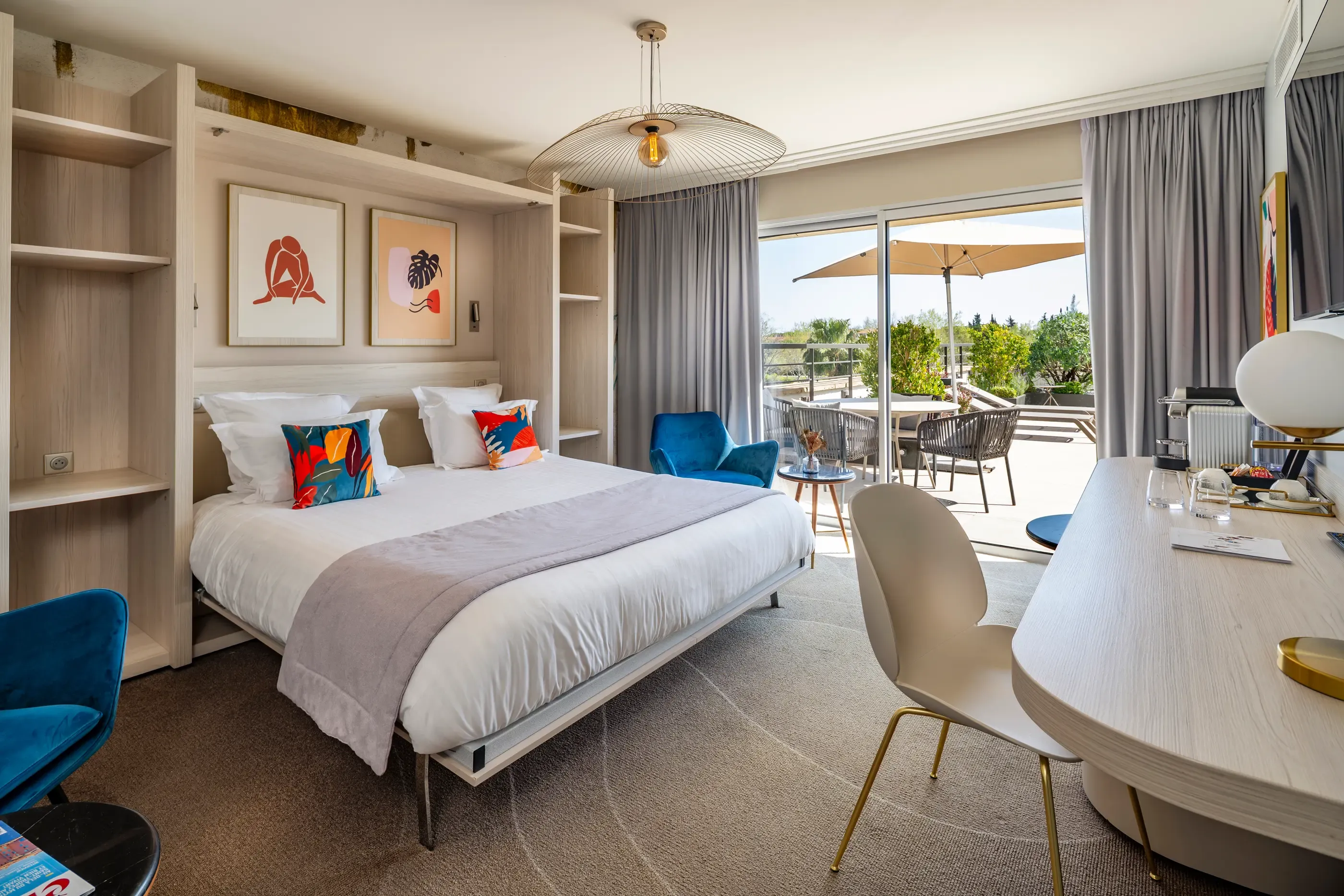 Chambre d'hôtel moderne avec un lit double, une table de travail, un fauteuil bleu et une vue sur une terrasse extérieure avec des tables et des chaises