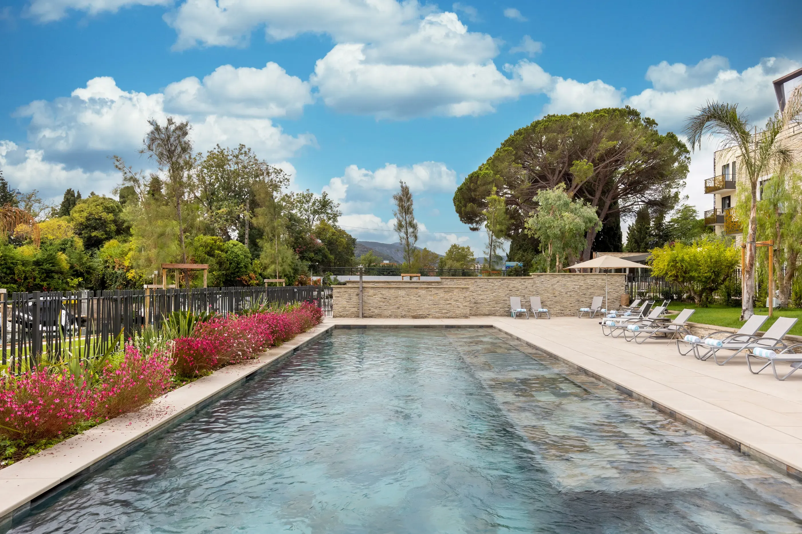 Piscine rectangulaire entourée de chaises longues et de végétation, avec un ciel partiellement nuageux en arrière-plan