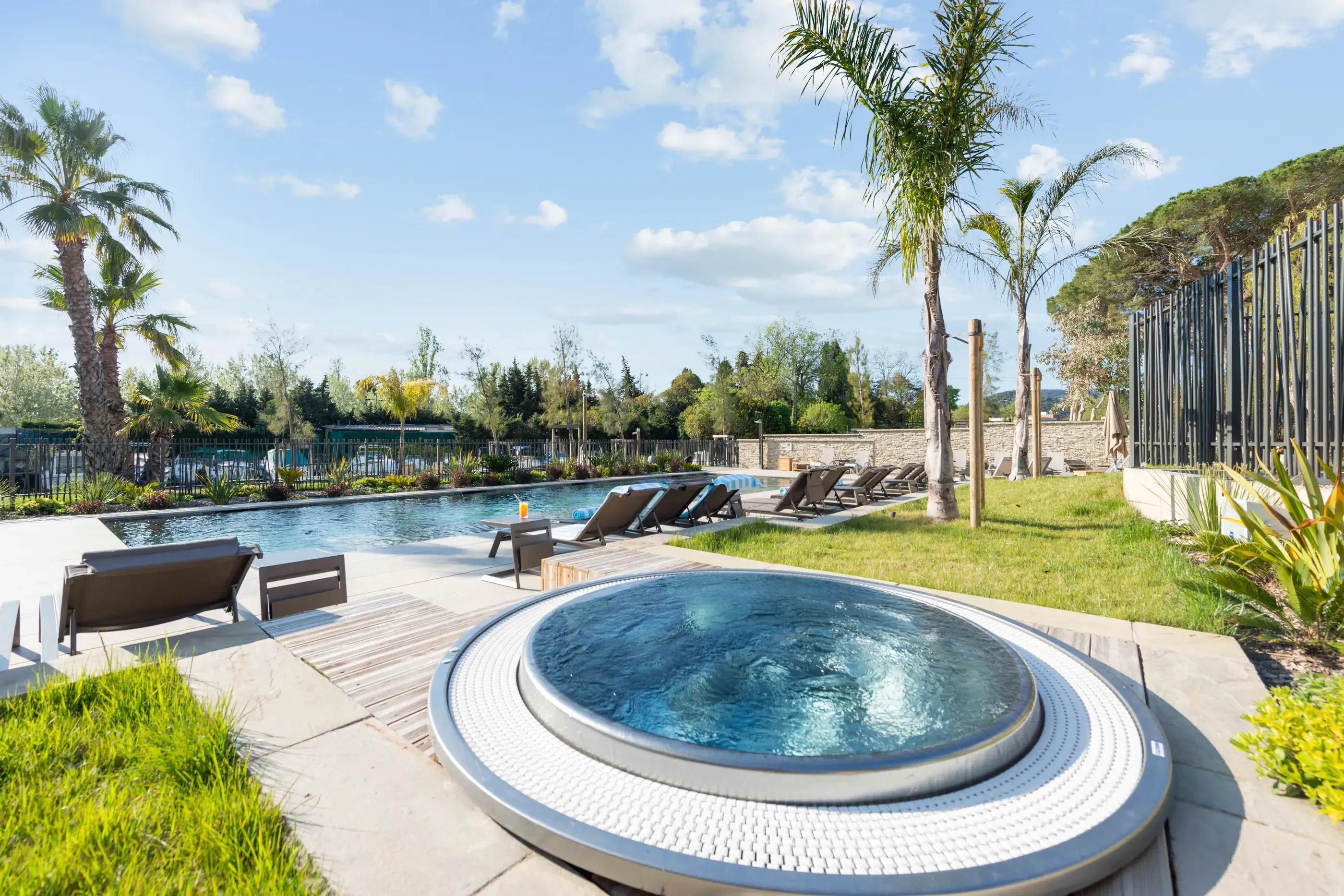 Piscine extérieure avec transats et un jacuzzi circulaire entouré de gazon et de palmiers sous un ciel partiellement nuageux