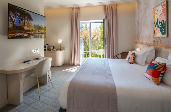 Chambre d'hôtel avec un lit, un bureau, une télévision, une porte-fenêtre donnant sur un balcon et des œuvres d'art aux murs