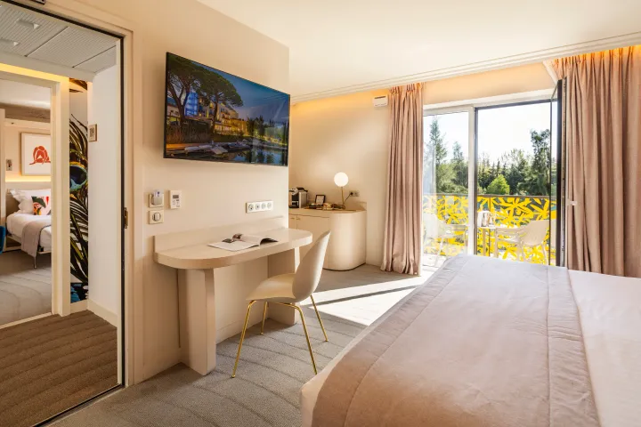 Chambre d'hôtel avec un lit, un bureau, une télévision et une porte-fenêtre donnant sur un balcon avec vue sur des arbres
