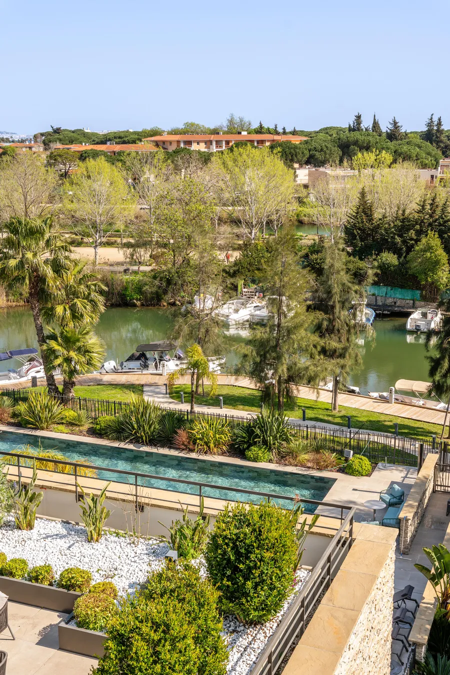 Vue d'un jardin avec piscine, bordé d'arbres et de plantes, donnant sur un plan d'eau avec des bateaux amarrés et des maisons en arrière-plan
