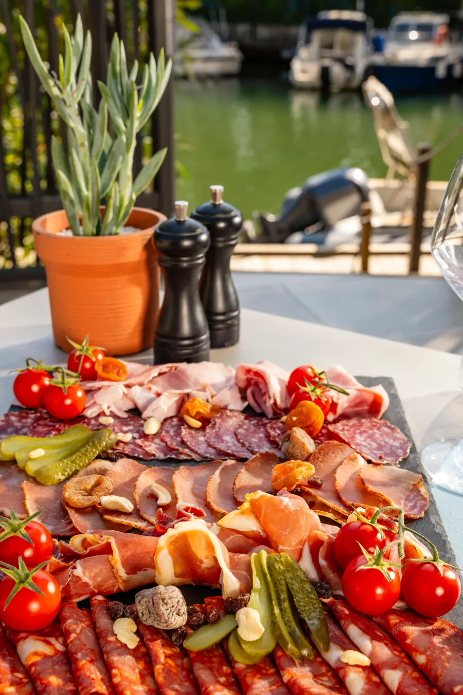 Un plateau de charcuterie avec différentes tranches de viandes, des tomates cerises, des cornichons et des noix, posé sur une table en extérieur près d'un plan d'eau