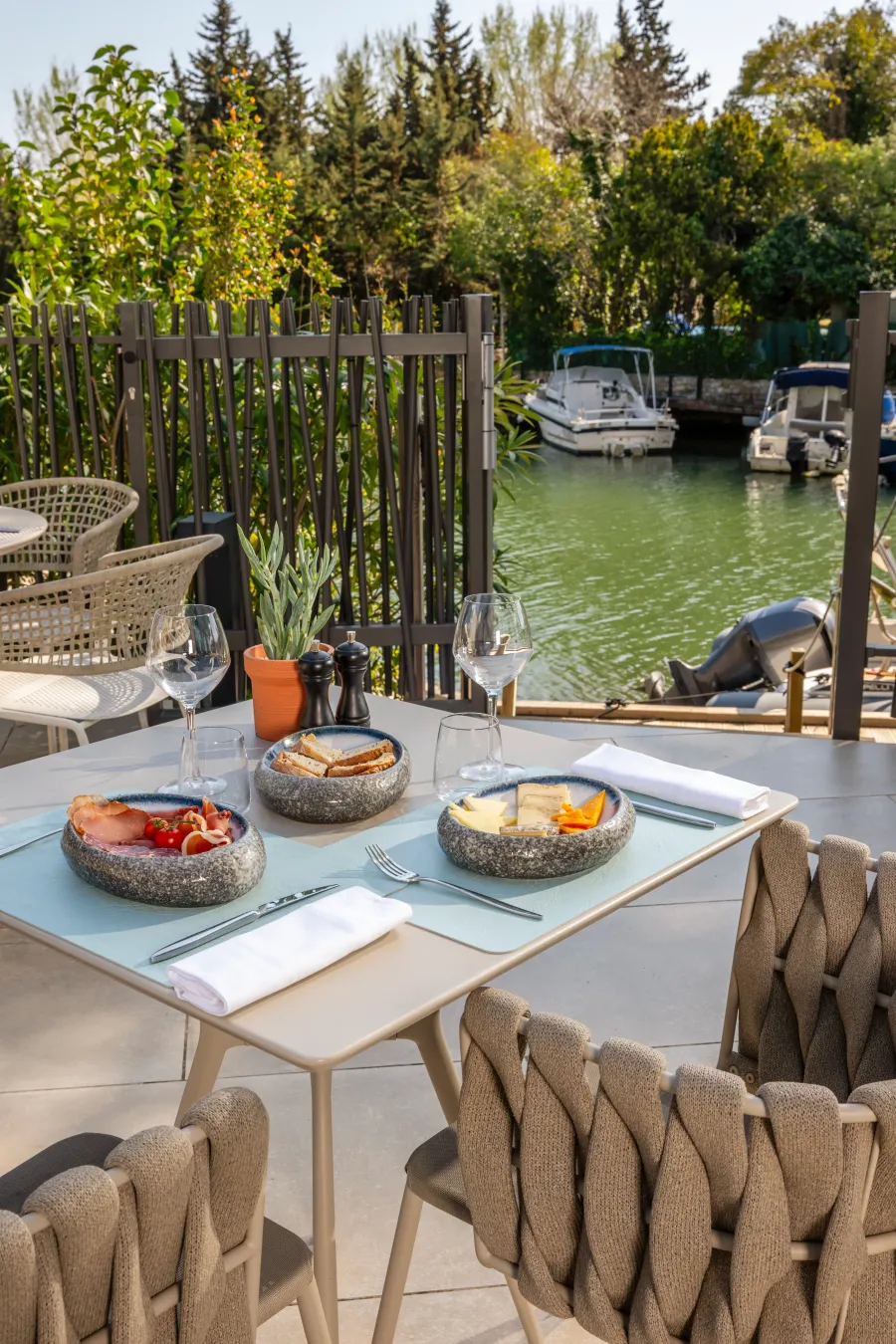 Table dressée en extérieur avec des plats, des verres et des couverts, face à un canal avec des bateaux amarrés et des arbres en arrière-plan