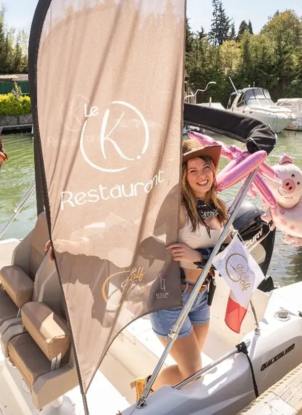 Une femme sourit sur un bateau avec une enseigne "Le K Restaurant" et un drapeau à l'arrière-plan